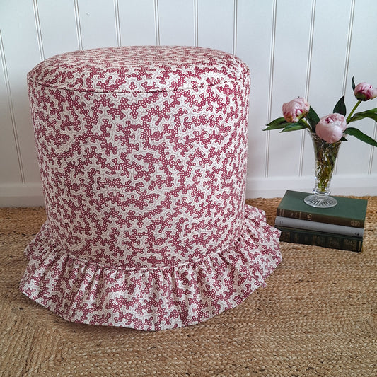 A round upholstered stool with a pink and white floral pattern and a ruffled skirt base, styled on a woven jute rug next to a vase of pink flowers and books against a white paneled wall.