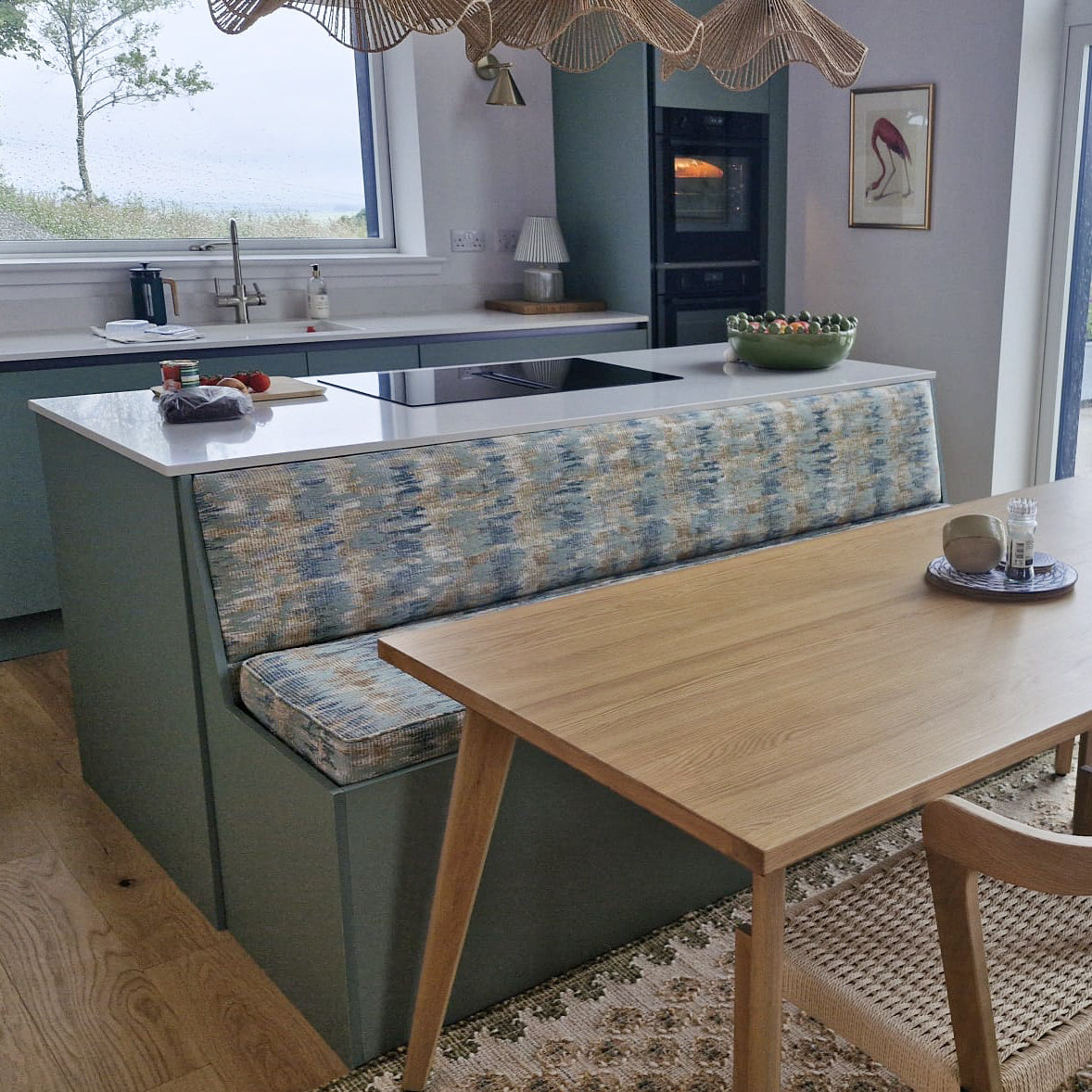 A modern kitchen island with a built-in bench seat upholstered in a blue and beige patterned fabric, set in a bright kitchen with a wooden dining table and chairs.